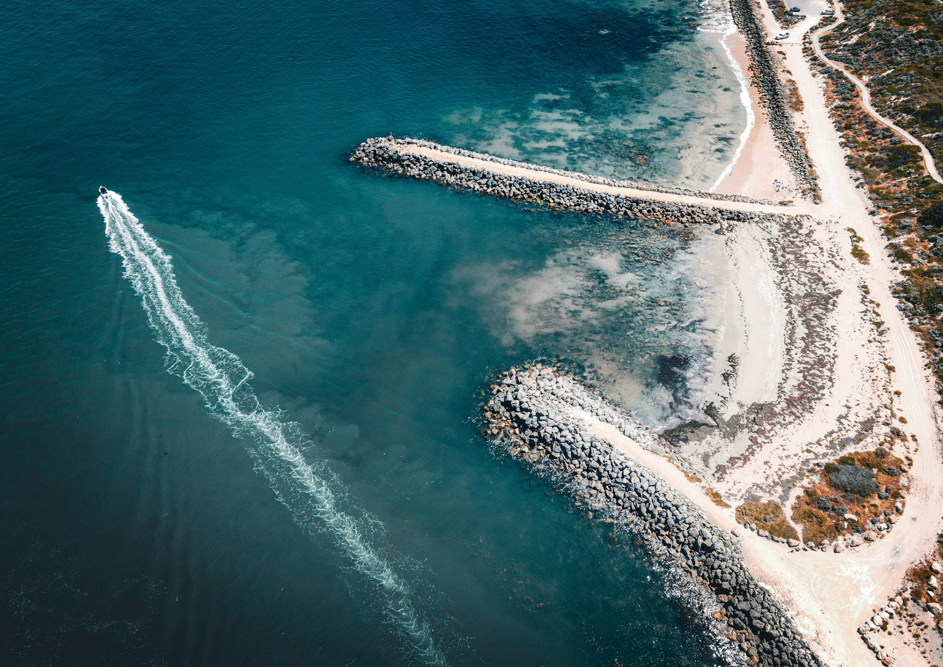 An aerial view shows a speedboat leaving a white wake as it travels through blue ocean water, heading towards a sandy coastline.
