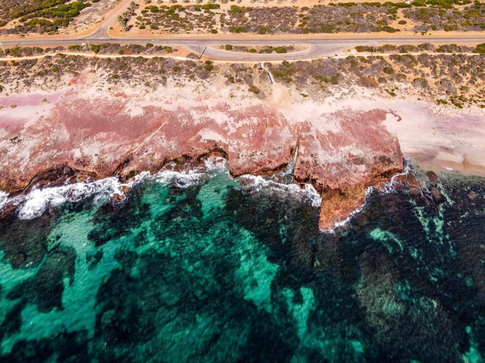 An aerial view showcases the rugged red-rock coastline and turquoise ocean waves. The serene landscape features crashing waves and a winding road.