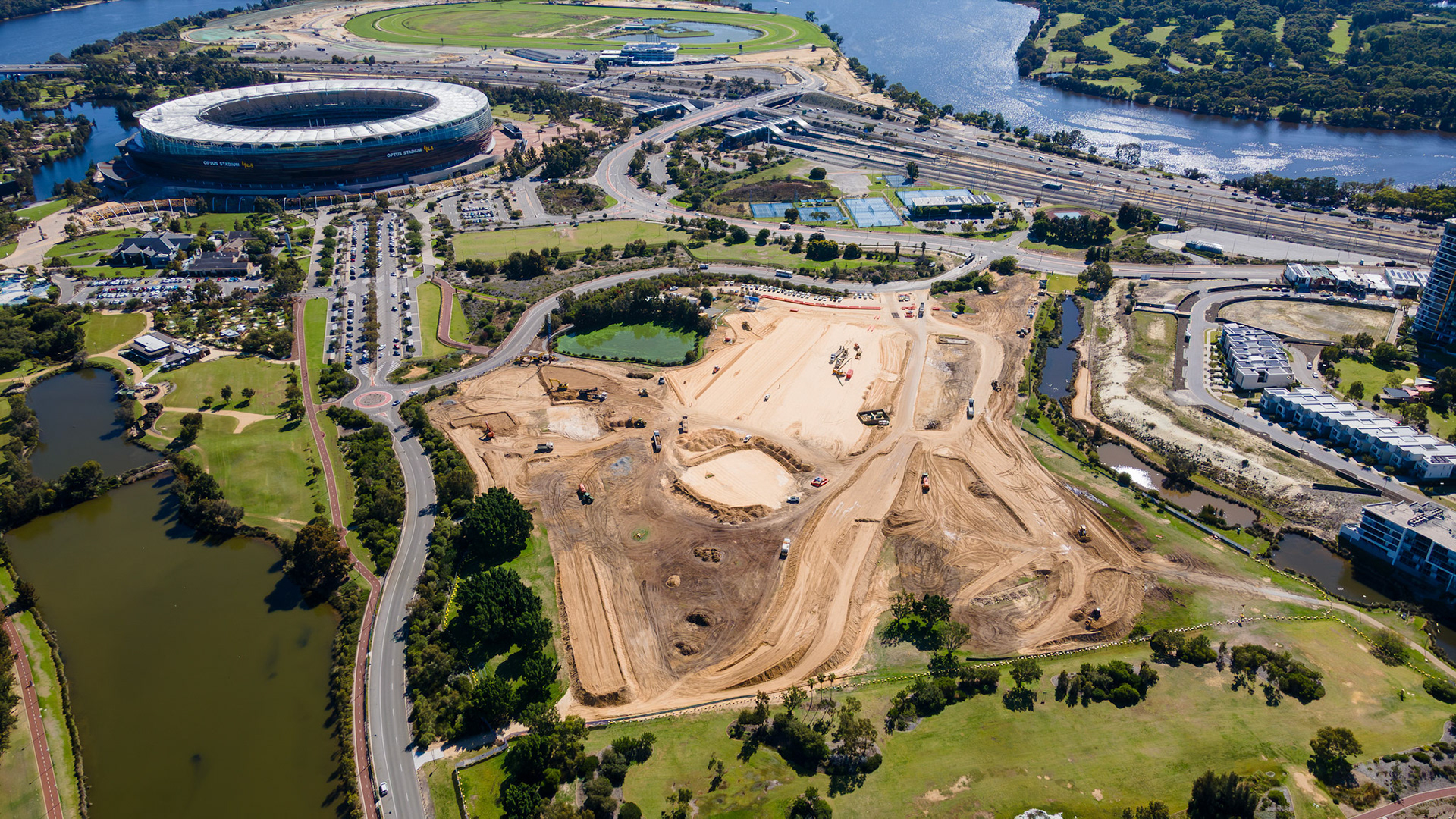 A large construction site unfolds near a vibrant stadium, surrounded by lush greenery and a winding river. Machines are busy reshaping the earth, marking progress in urban development.