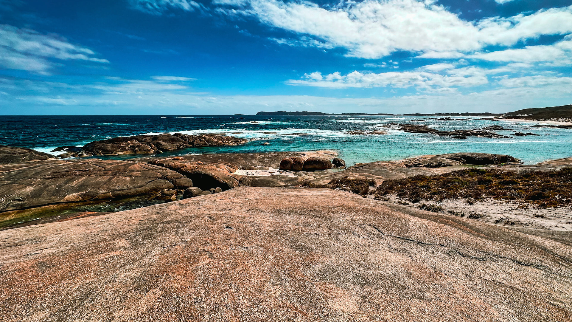 A rugged coastline meets the vast ocean under a bright blue sky. Waves crash against dark rocks, creating a serene seascape.