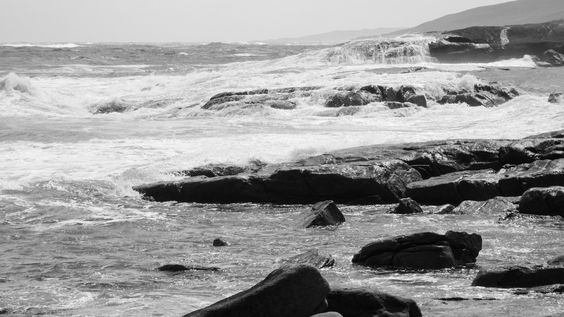 A black and white image capturing the dynamic interaction of ocean waves crashing onto a rocky shoreline. The shoreline is rugged with large rocks, and the waves create a dramatic, foamy texture against the smooth surface of the water. In the distance, a faint silhouette of coastal hills enhances the scenic beauty and adds depth to the composition. The photograph conveys a sense of tranquility and raw natural power.