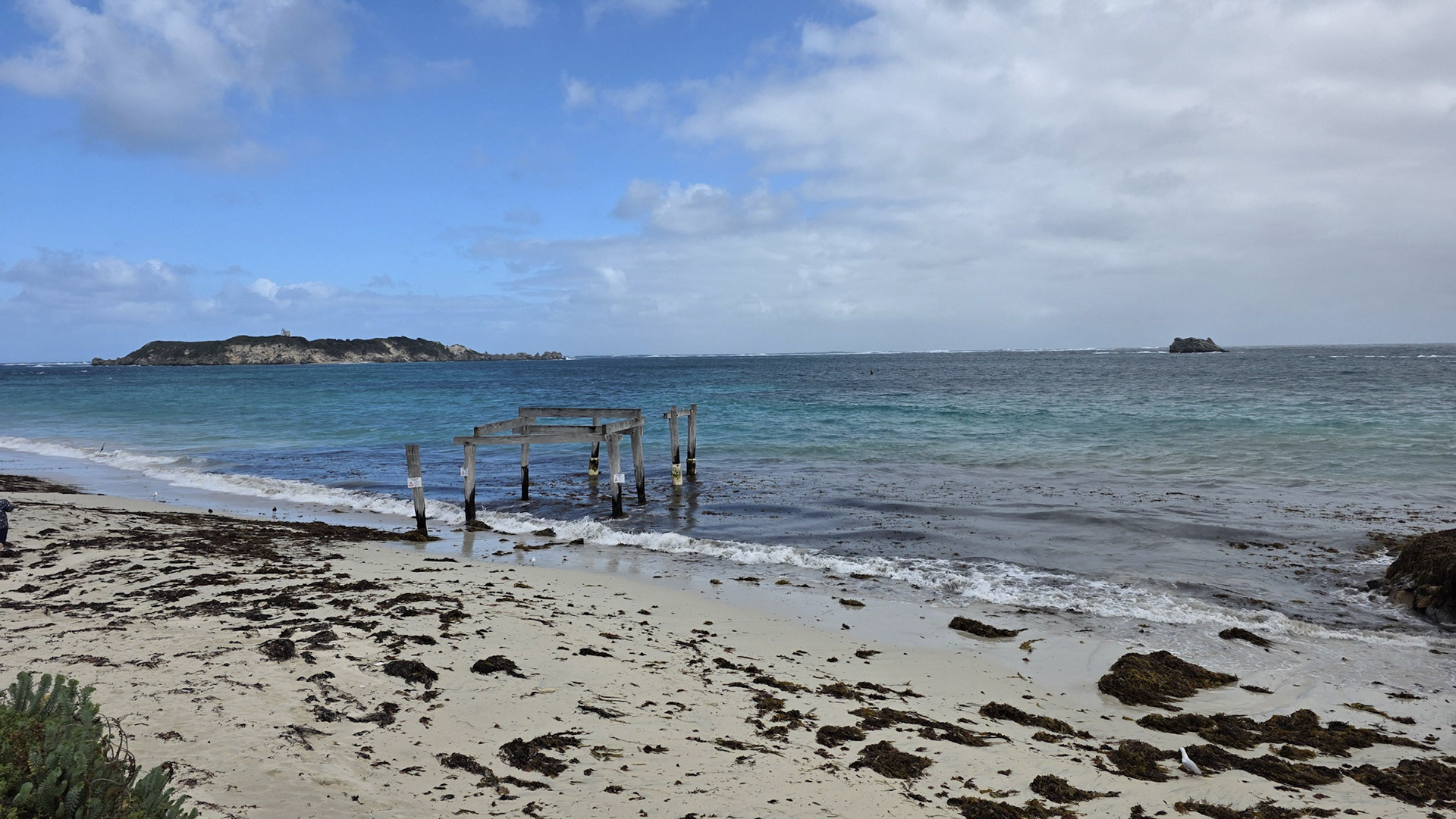Scenic view of a peaceful beach with gentle waves lapping against the shore, capturing an old, weathered pier jutting into the turquoise waters. Sparse clouds dot the blue sky, and distant islands enhance the serene ocean landscape. Seaweed scattered across the sandy beach adds natural texture to the coastal scene.