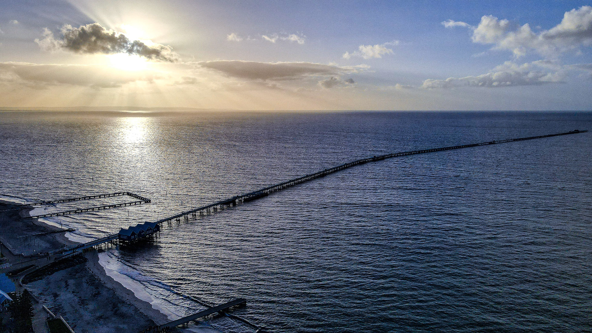 Aerial view of a long pier extending into a serene ocean as the sun sets on the horizon. Sunrays peek through scattered clouds, casting shimmering reflections on the water. This tranquil seascape captures the peaceful beauty of nature and maritime structures, ideal for themes of travel, relaxation, and coastal living.