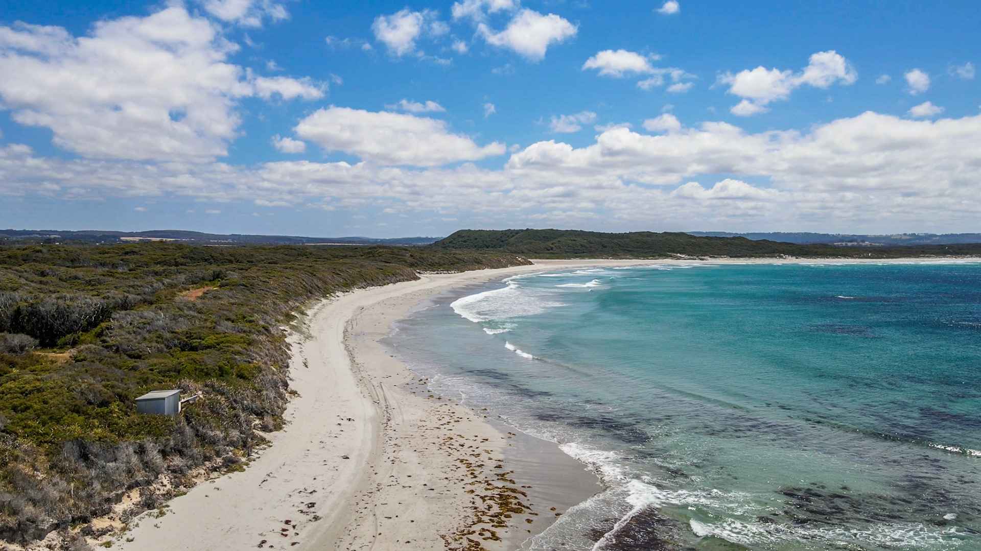 An aerial perspective showcases a tranquil beach meeting the ocean, surrounded by lush vegetation and a bright blue sky with clouds.