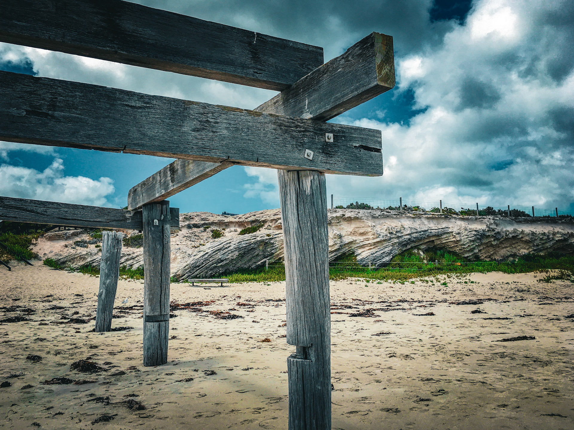 Weathered wooden posts and beams stand on a sandy beach, overlooking dunes and a dramatic sky. This scene reflects nature's resilience.