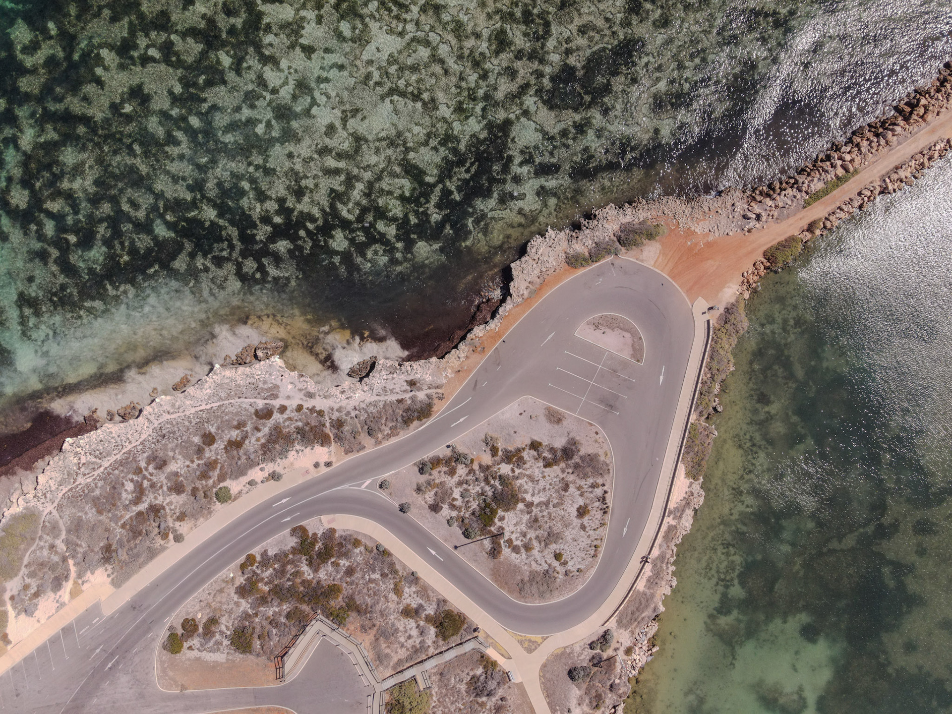 An aerial view showcases a winding coastal road meeting the ocean, surrounded by serene scrubland and vibrant waters.