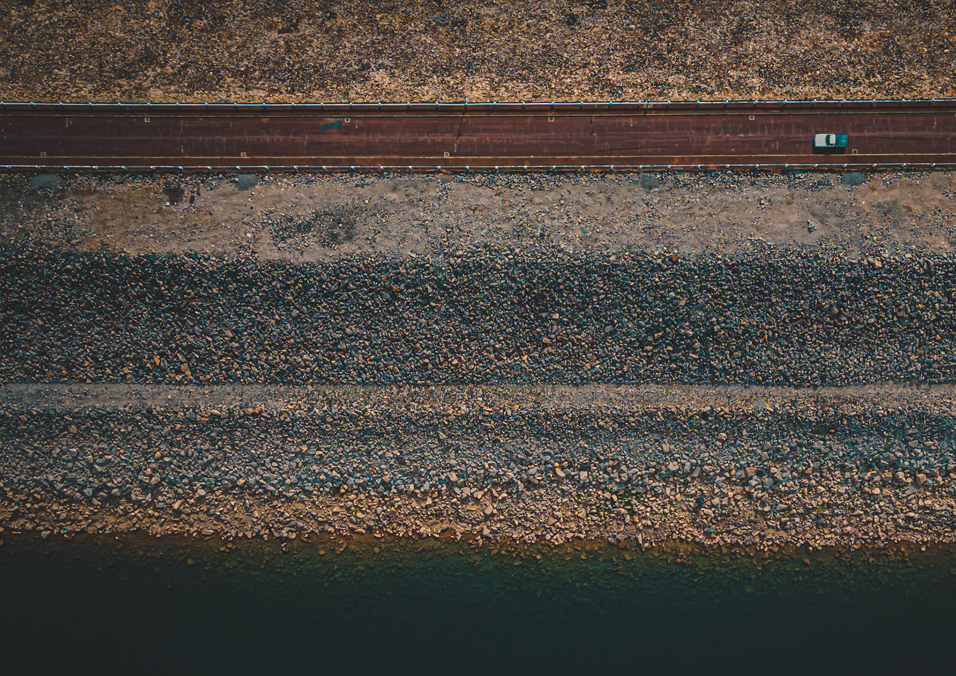 A car drives along a road next to a rocky coastline. The water is visible alongside the pebbly shore. Light reflects on the water during the day.