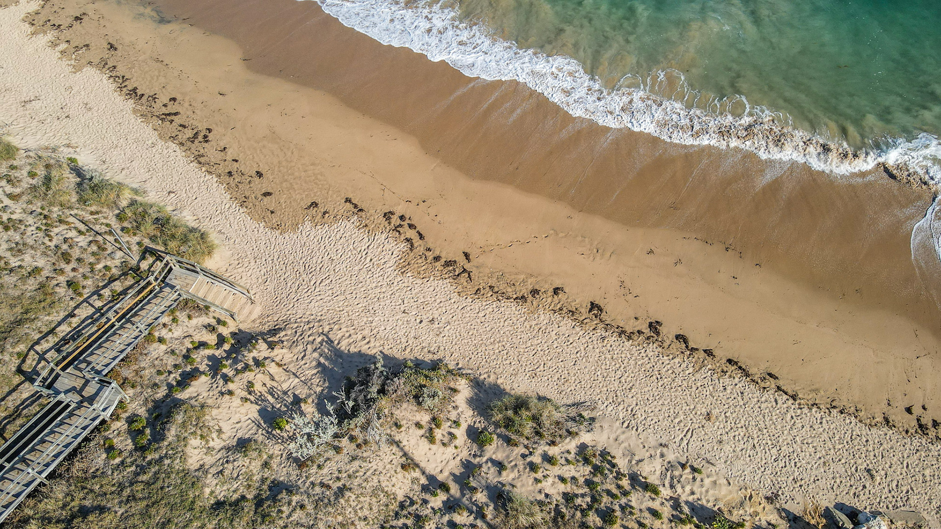 An aerial view showcases a tranquil beach scene with gentle waves, a wooden boardwalk, and the natural beauty of the coastline.