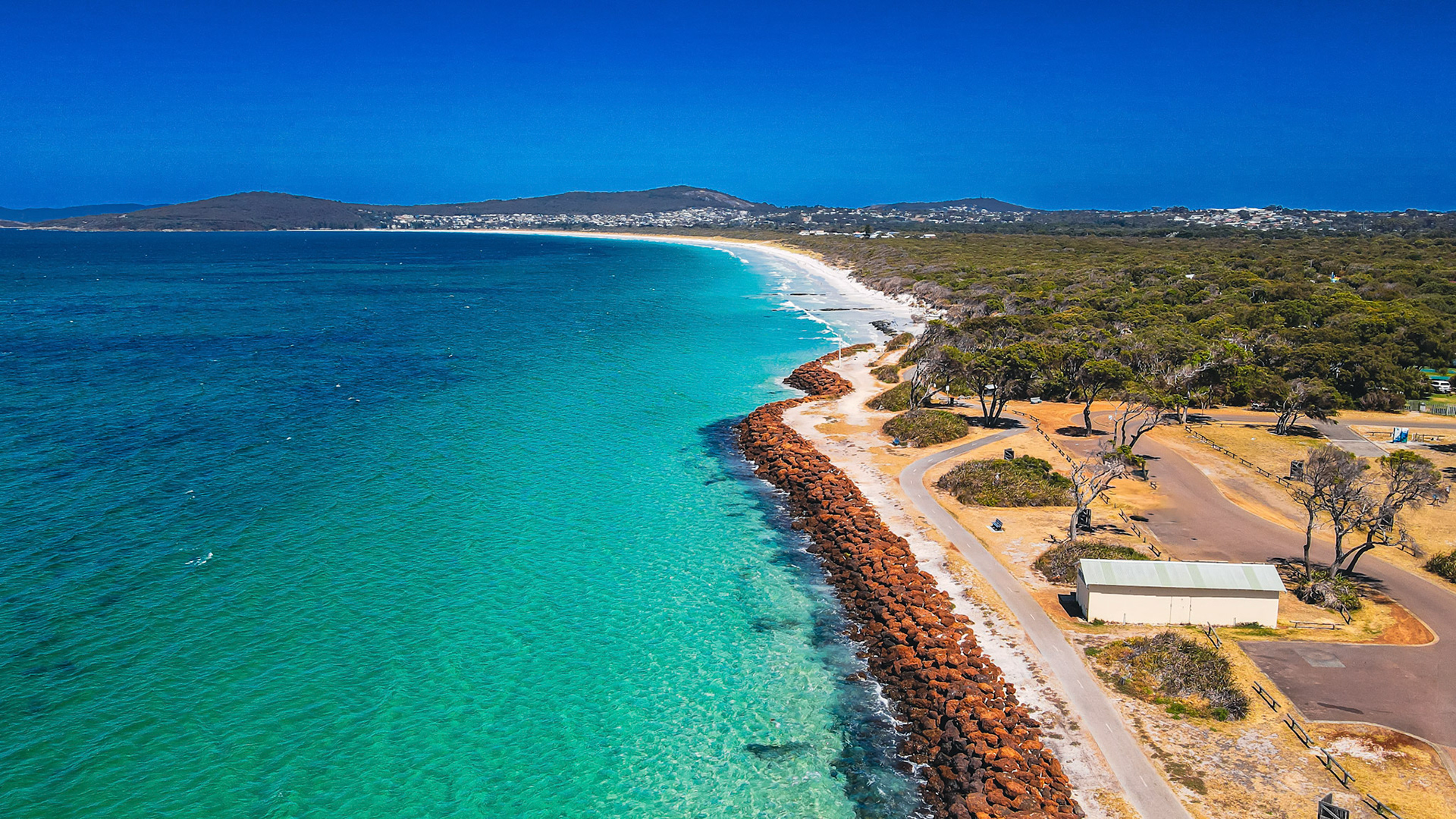 An aerial view showcases a coastal landscape with turquoise waters, a sandy beach, and a rock wall, evoking serenity and natural beauty.