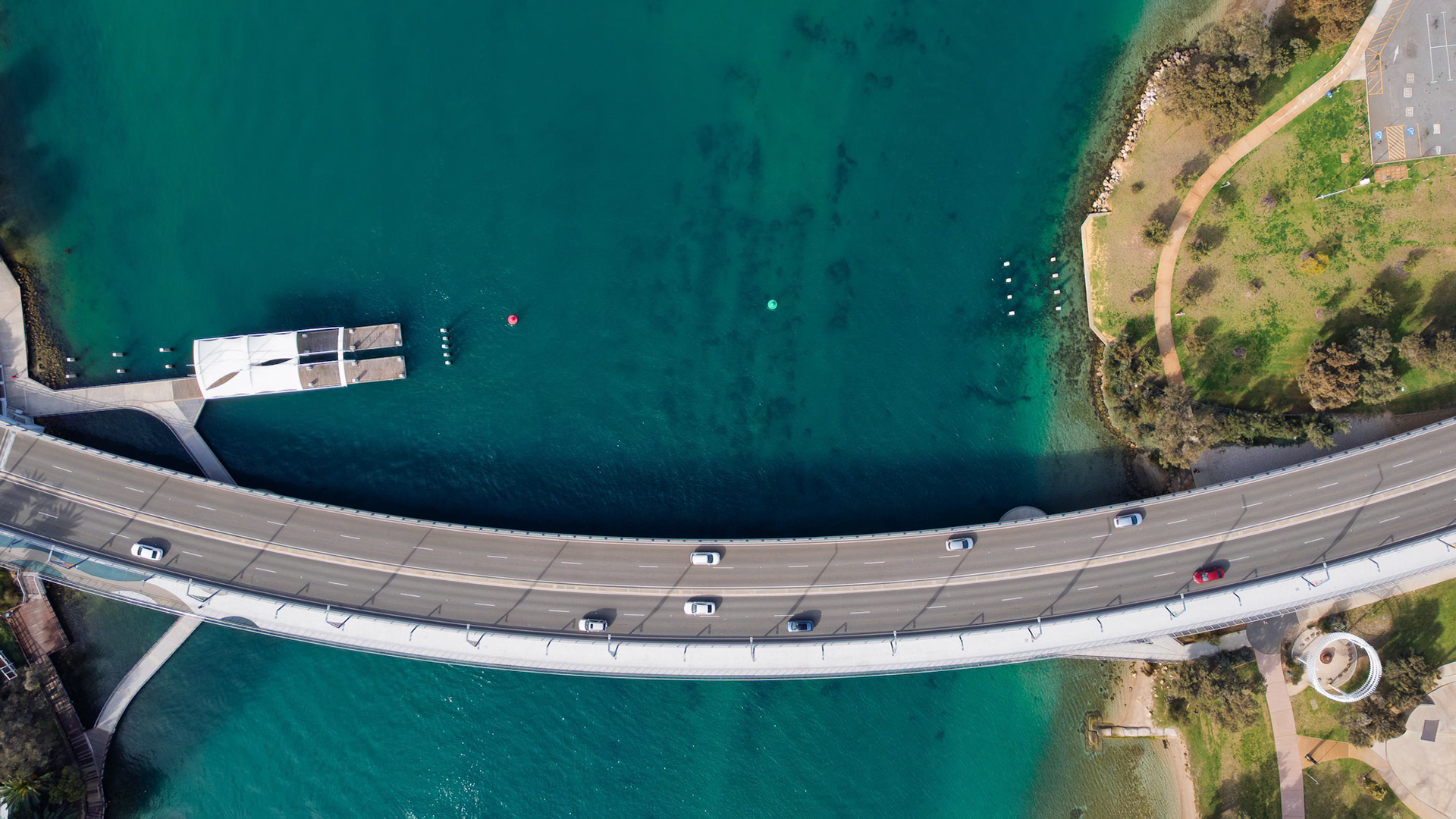 This stunning aerial shot captures a modern curved bridge that spans across a vibrant turquoise body of water, creating a scenic view. The photo highlights the smooth asphalt road, alongside vehicles in motion, surrounded by lush greenery and a peaceful marina. The image effectively showcases both nature and infrastructure, making it ideal for various themes such as travel, engineering, and environmental studies. The contrast between the deep blue water and the light-coloured bridge adds depth to the composition, enhancing its visual appeal for marketing and multimedia projects.