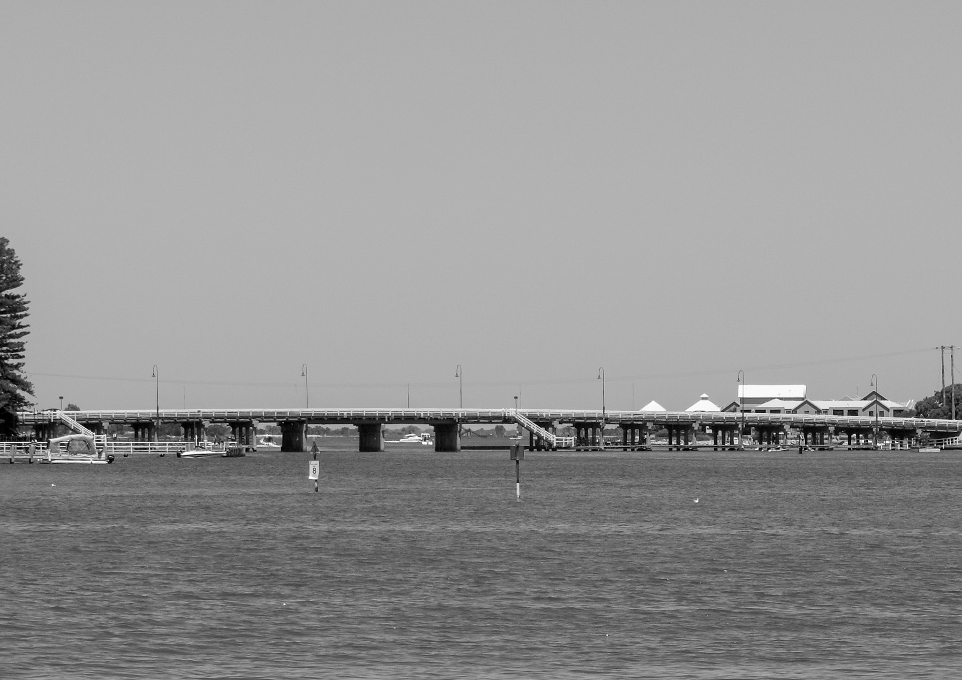 A black-and-white view of a low bridge stretching across calm estuary waters. Small boats and jetties sit along the foreshore, and a stairway descends from the bridge to the water. Lamp posts line the span, while trees and low buildings frame the shoreline beneath a clear, open sky, creating a quiet, coastal atmosphere.