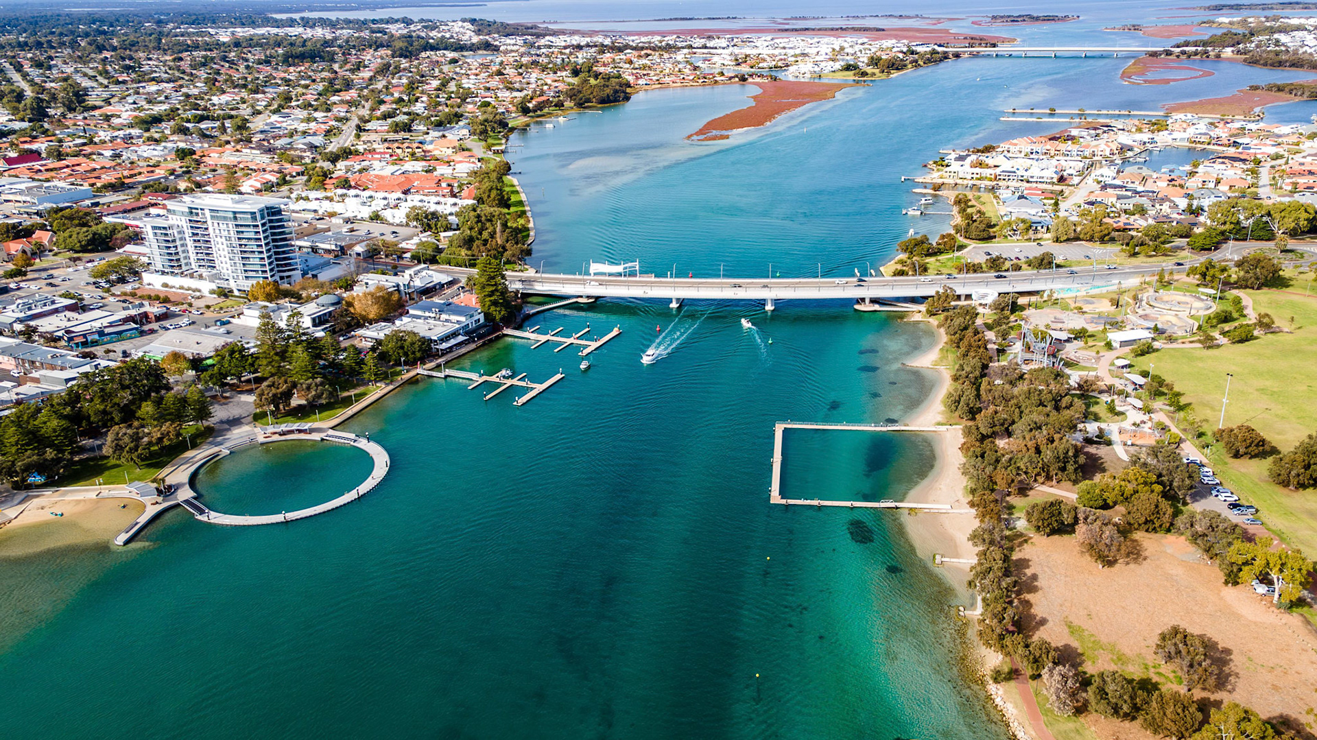 Stunning aerial shot showcasing a vibrant waterfront area. This image captures the essence of a picturesque neighborhood with inviting waterways, local marinas, and waterfront parks. The scene includes residential buildings, a pedestrian beach area, and recreational facilities, surrounded by lush greenery. Ideal for real estate, tourism, and leisure-oriented projects, this stock image features clear turquoise waters and a well-maintained marina, making it a perfect representation of urban coastal living.
