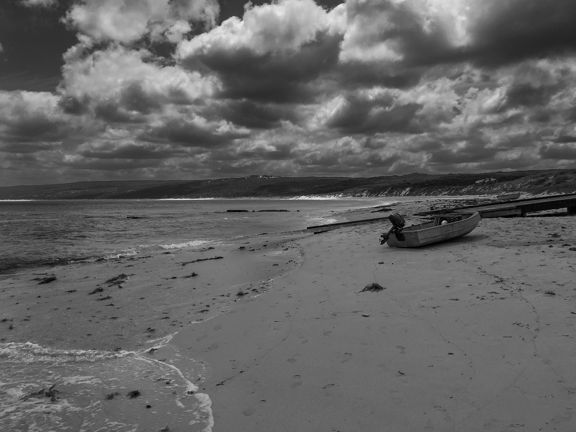 A weathered rowboat sits on the sandy beach, surrounded by ocean waves and a cloudy sky. This black and white scene evokes solitude.
