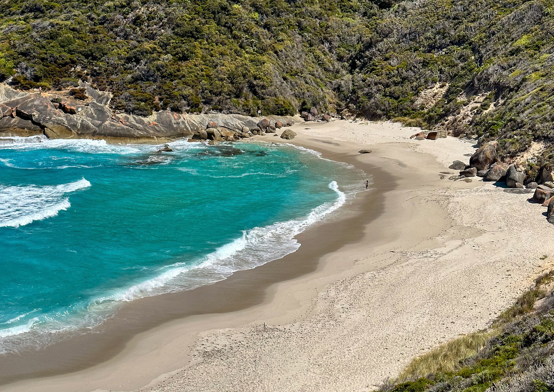 A solitary figure walks along a secluded beach, surrounded by cliffs and lush vegetation, evoking tranquility and peace.