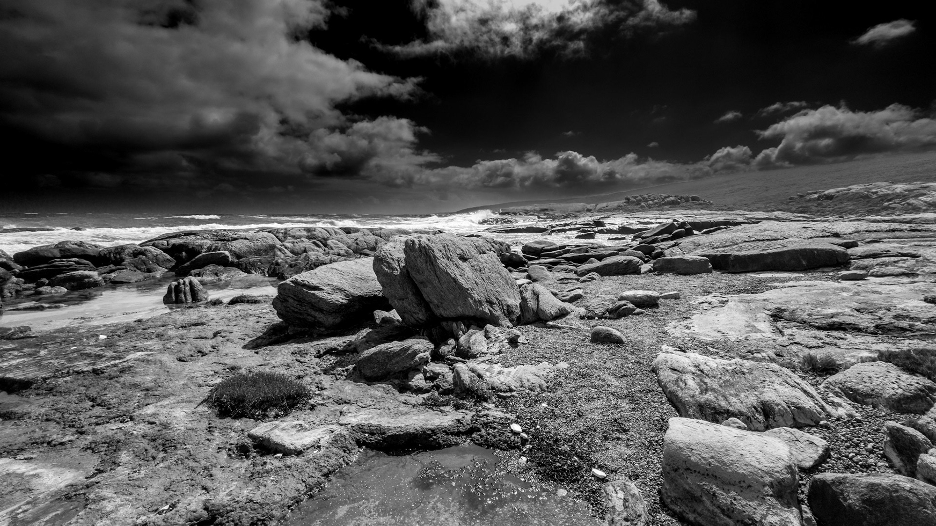 A striking black and white image capturing a rugged rocky coast under a dramatic sky. The contrast between the textured stones and the ominous clouds creates a powerful and moody atmosphere, perfect for conveying themes of nature's raw beauty and strength.