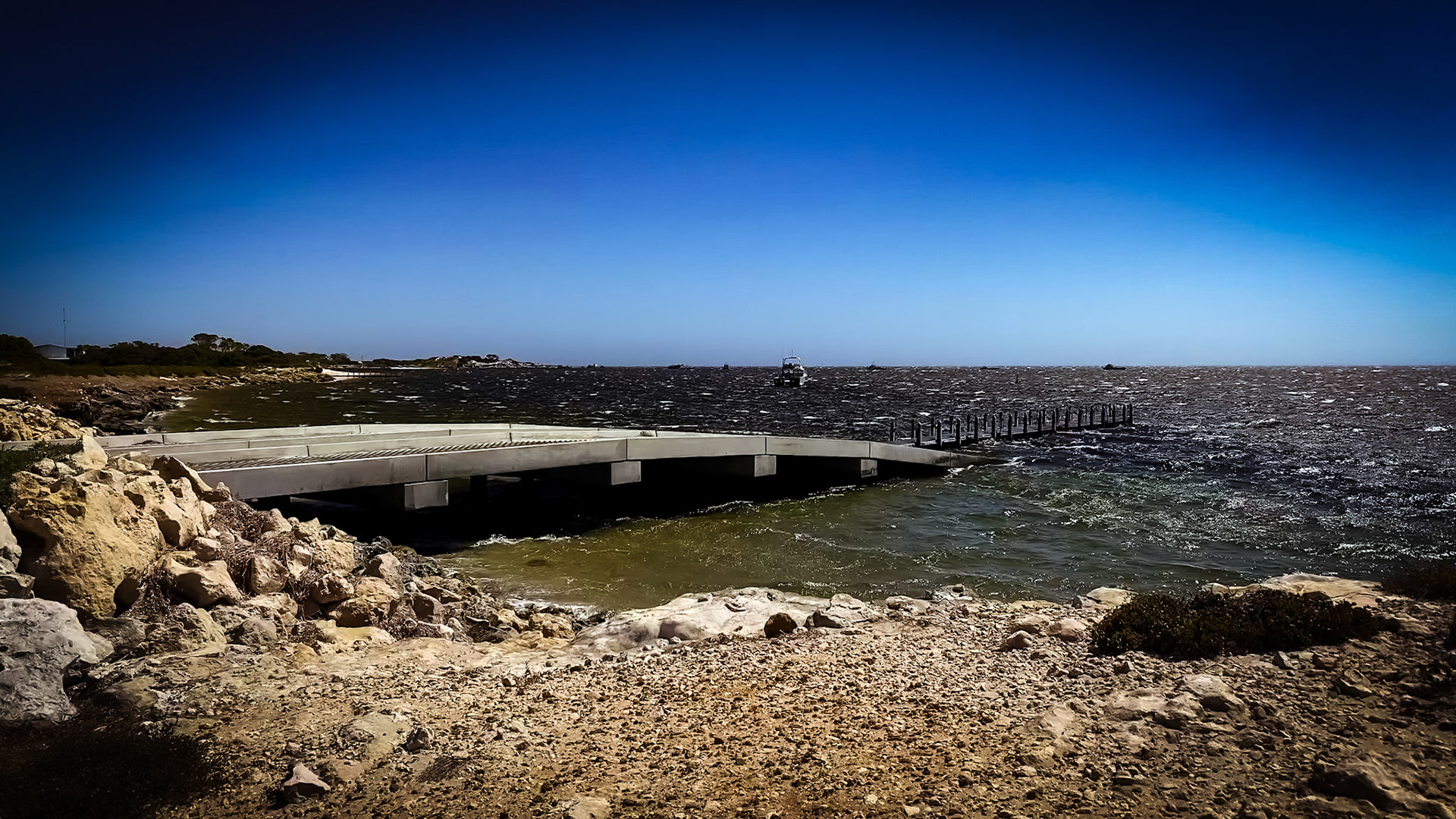 A low concrete boat ramp and narrow wooden jetty extend into choppy, dark water under a deep blue sky. A small boat sits on the horizon while the foreground shows sunlit, weathered rocks and sparse coastal vegetation. Bright daylight highlights texture in the stones and rippling waves, creating a stark, windswept seascape.