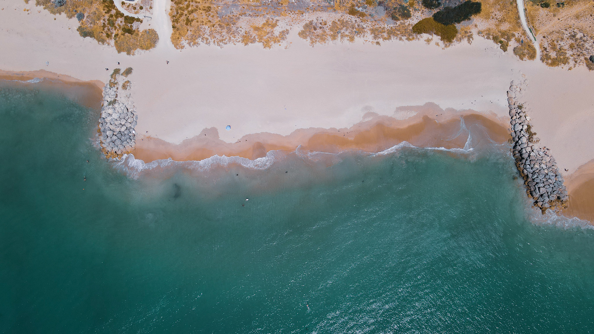 A top-down drone view of a quiet beach where teal water laps onto pale sand. A warm, rust-tinted swash line traces the shore, while two stone groynes on either side shape the small cove. A few scattered beachgoers and a lone blue umbrella dot the wide, open sand. Behind the beach, low dunes and a winding path add subtle texture to the coastal landscape.