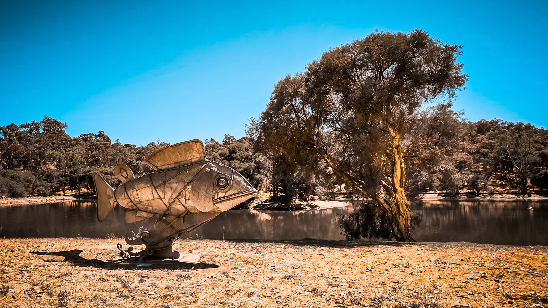 A large fish sculpture made of metal stands on a sandy bank near water, surrounded by trees and a bright blue sky.