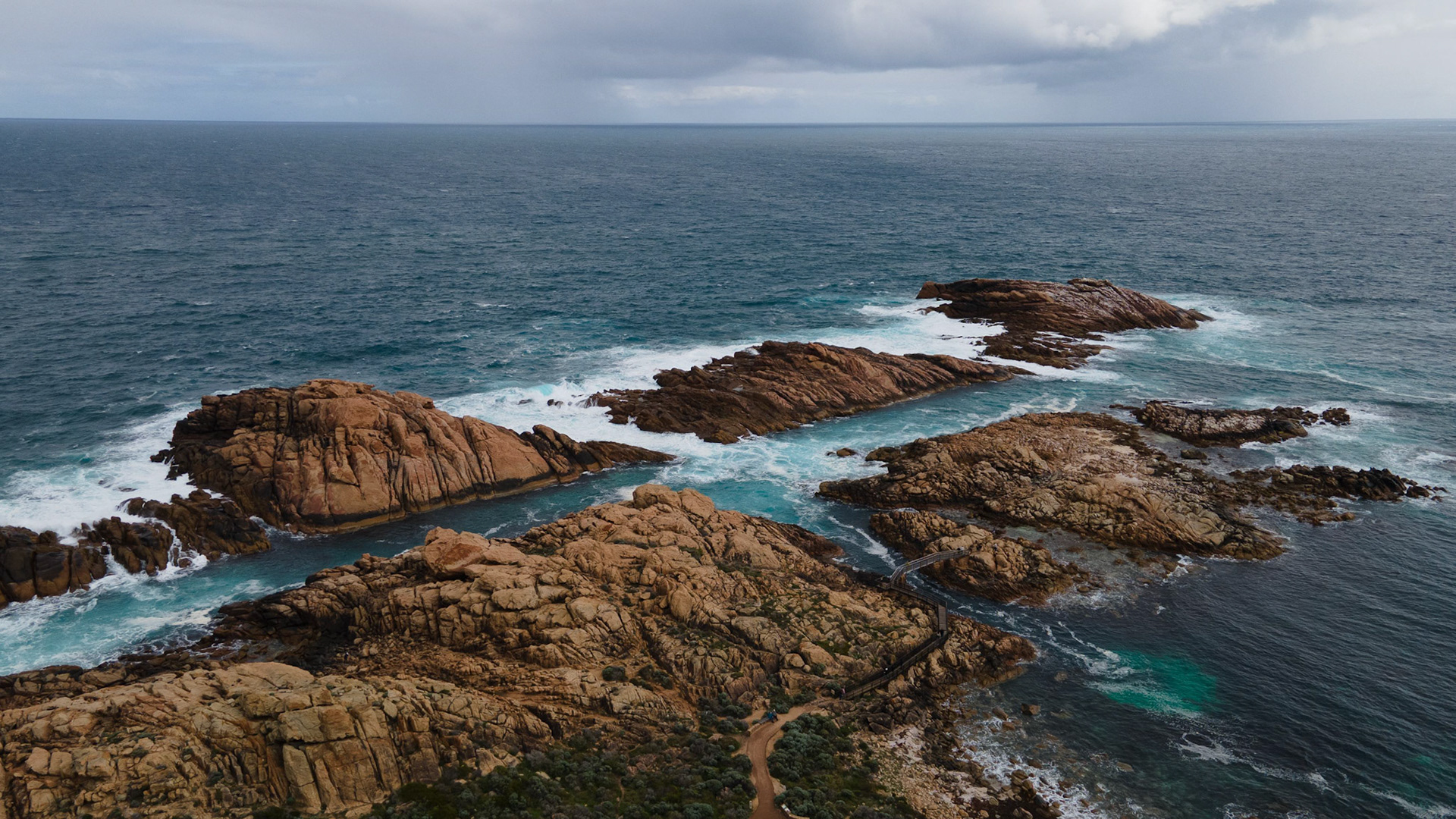 A stunning aerial shot captures the dramatic coastline featuring rugged rocky formations and the vibrant blue ocean waves crashing against the rocks. The scene is enveloped by overcast skies, adding a moody and atmospheric touch. This natural landscape showcases the raw beauty of nature with its intricate rock patterns, expansive ocean, and the meeting of land and sea.