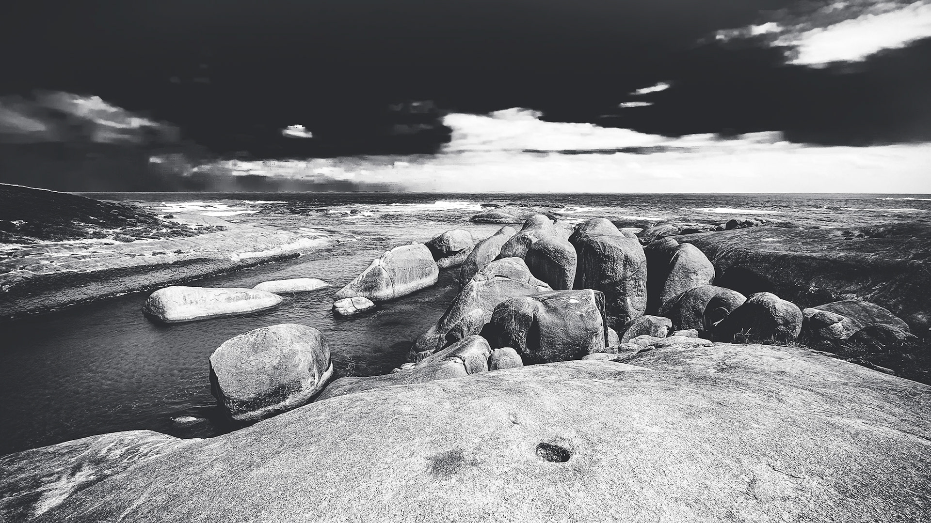 A dramatic black and white landscape features rugged boulders along a serene coastline, reflecting the moody sky and water.