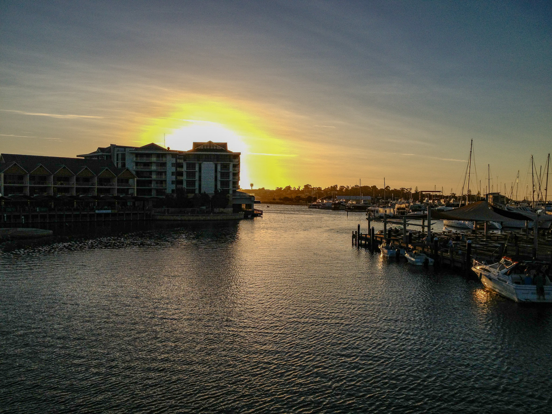 The image captures a serene marina scene at sunset in Mandurah, Western Australia. The golden sun is setting behind a modern waterfront building, casting a warm glow over the water. Several boats are moored at the wooden docks to the right, while the calm water gently reflects the fading light. The sky transitions from a golden hue near the horizon to deep blue above, enhancing the tranquil and picturesque atmosphere of the harbor.