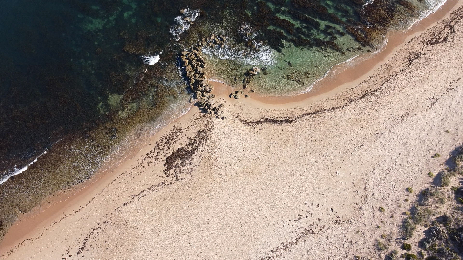 An aerial perspective showcases a serene beach where gentle waves meet the ocean. The tranquil scene highlights clear water over rocky outcrops.