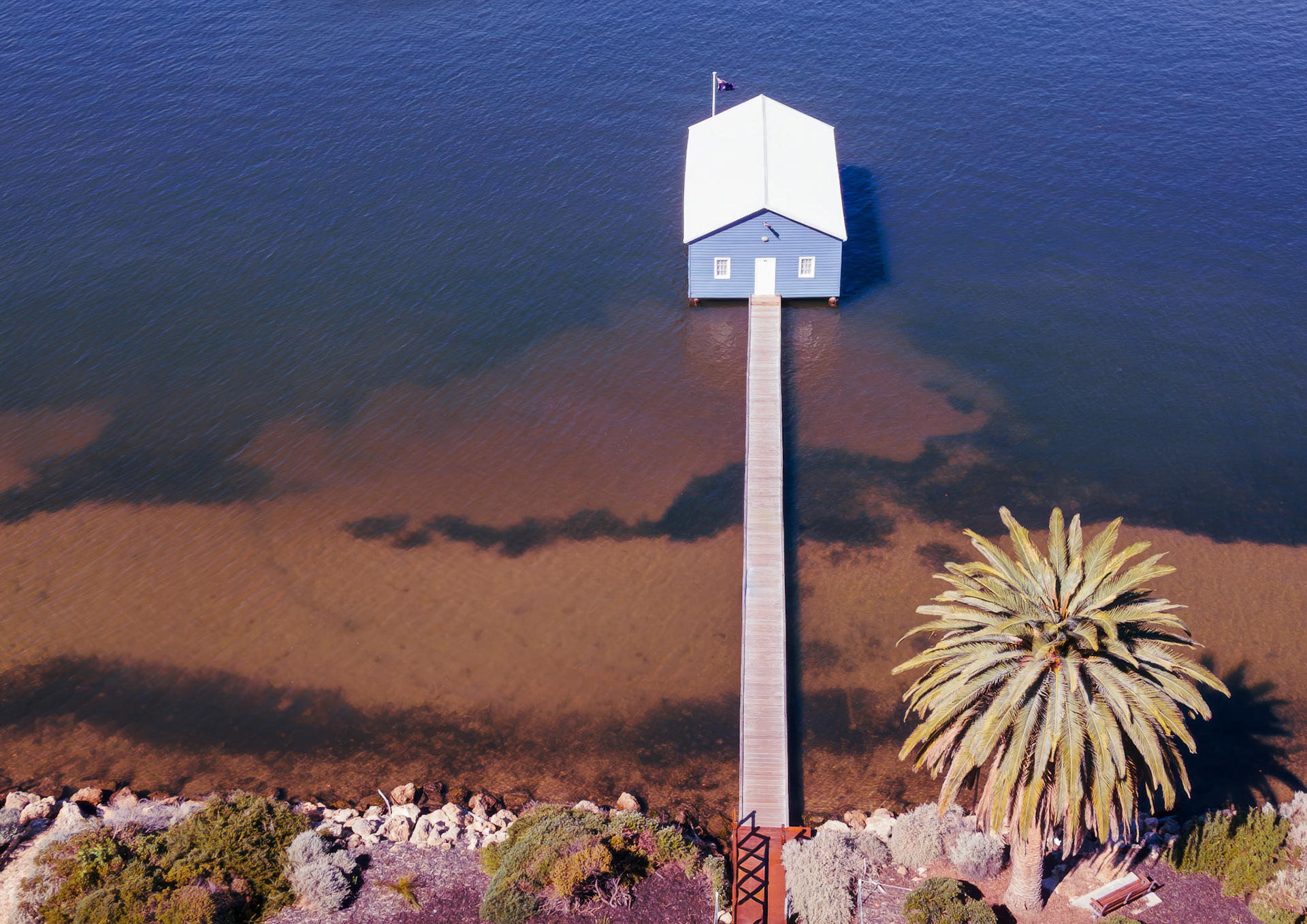 This tranquil scene captures a blue boathouse nestled on still waters, surrounded by lush palm trees and rocky shores. The golden light of sunset casts a beautiful glow over the landscape.