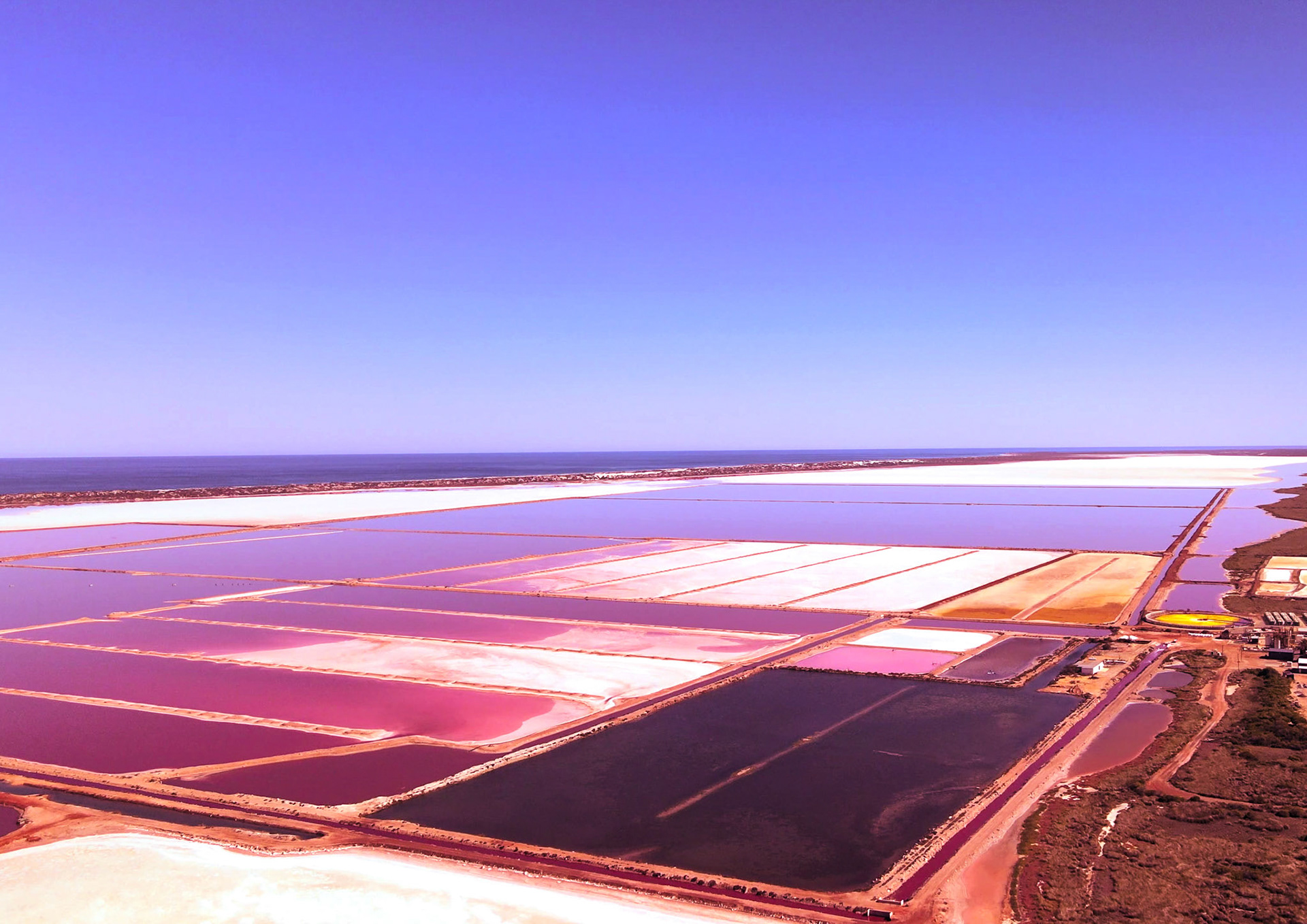 An aerial view showcases vast salt flats with pink water and an industrial facility. The landscape features numerous ponds and clear blue sky.