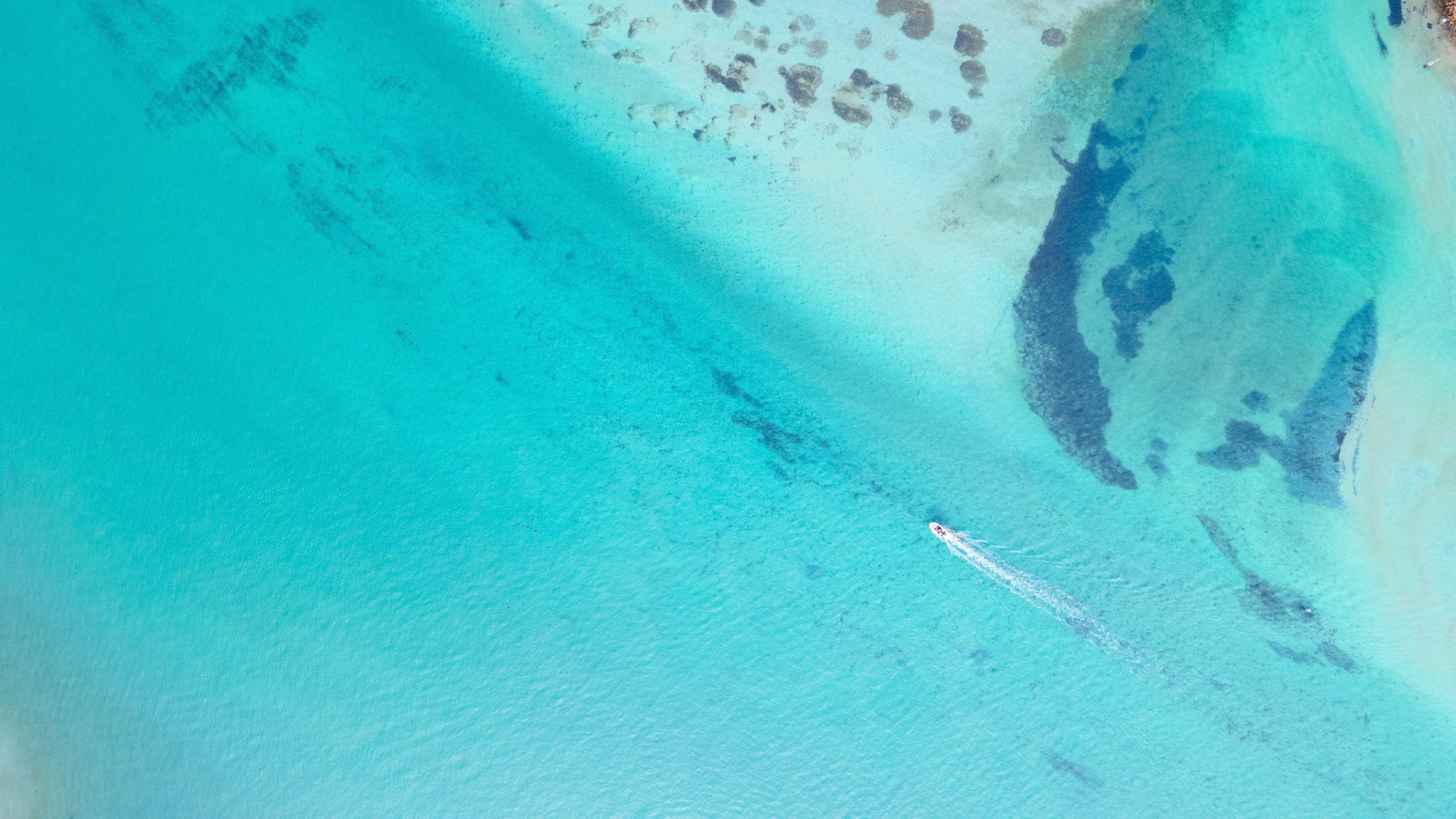 This stunning aerial photograph captures a speedboat gliding across vibrant turquoise waters, showcasing the beautiful contrast between the water and the underwater textures. The image highlights the serene and inviting nature of tropical destinations, making it ideal for travel blogs, marketing materials, or lifestyle publications. The top-down perspective encourages viewers to imagine themselves on an exciting adventure or relaxing getaway, making it a versatile image for various commercial applications.