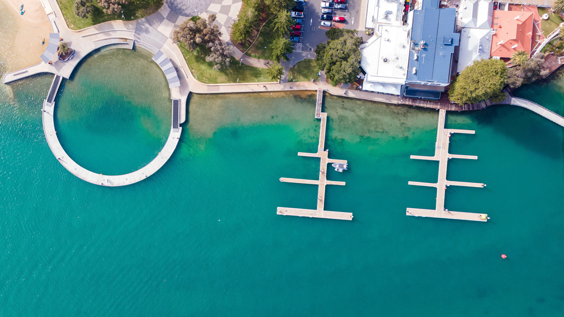 Stunning aerial shot capturing a marina with modern docks in a vibrant turquoise water setting. The image features well-maintained wooden docks with boats, surrounded by lush greenery and sandy beach areas. The clear blue water reflects the serene environment, making it ideal for vacation destination promotions, marina advertisements, or travel-related content.