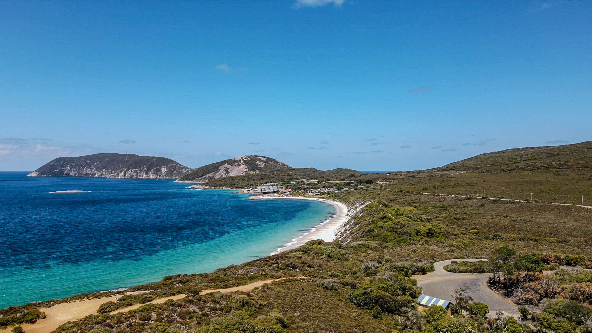 Aerial view of a sweeping crescent-shaped beach with bright white sand and vibrant turquoise water fading into deep blue. Low coastal scrub blankets rolling hills, a winding road traces the shoreline, and a small cluster of buildings sits near the water. Distant rocky headlands frame the bay beneath a clear, sunny sky.