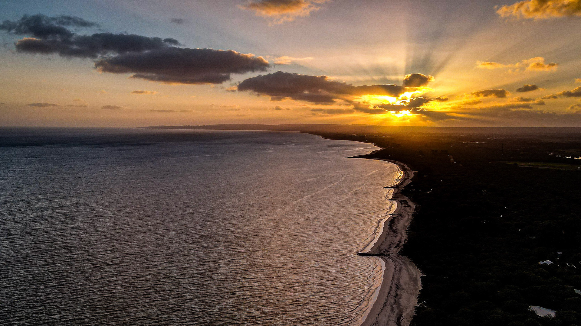 Stunning aerial photograph capturing the mesmerizing view of a golden sunset over a tranquil coastline. The sun's rays pierce through scattered clouds, casting a warm glow across the ocean and shoreline. The gentle waves ripple towards a pristine beach, creating a serene and picturesque landscape.