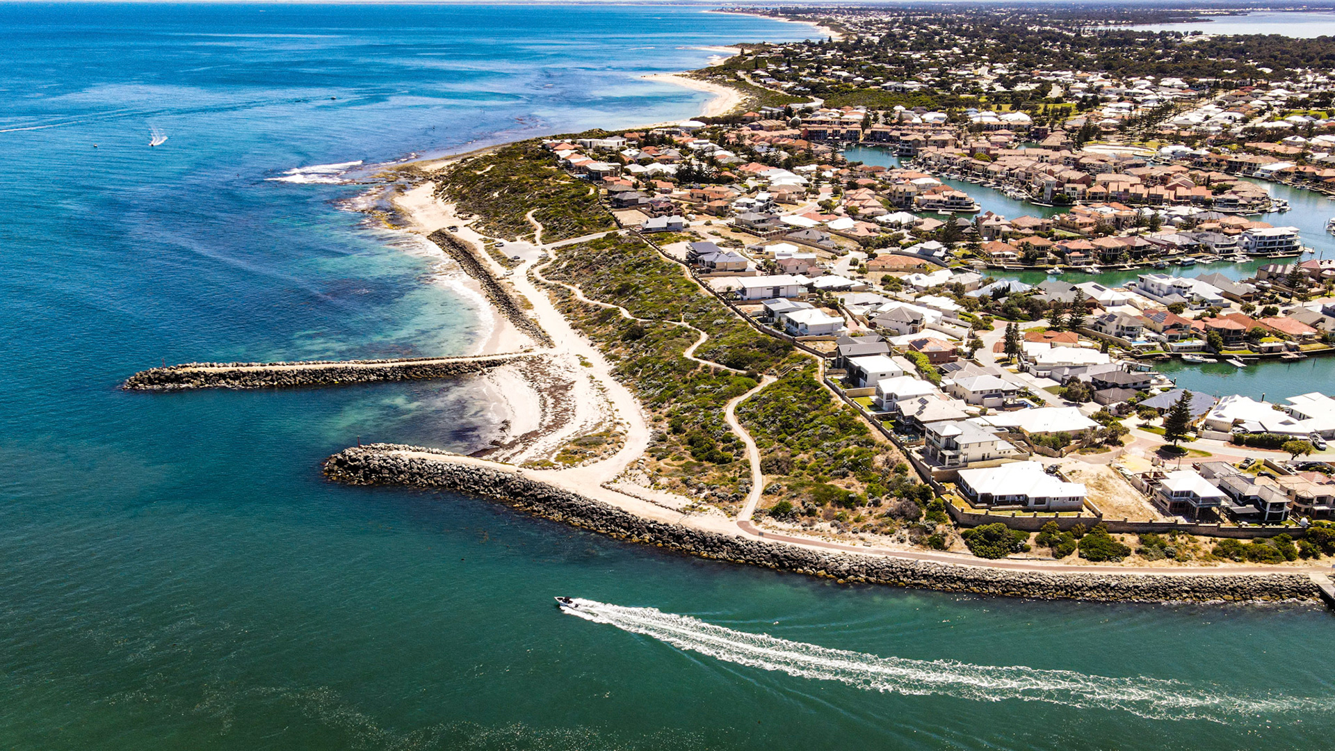 An aerial view shows a coastal town featuring waterfront homes and canals. A boat speeds across the turquoise ocean water.