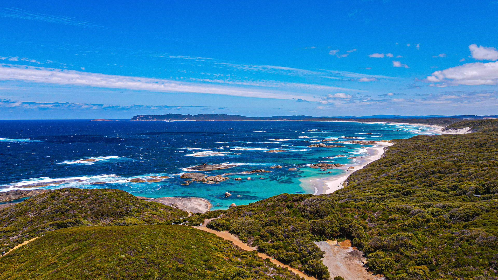 An aerial view showcases a rugged coastline with vibrant turquoise waters, rocky outcrops, and a sandy beach under a bright blue sky.