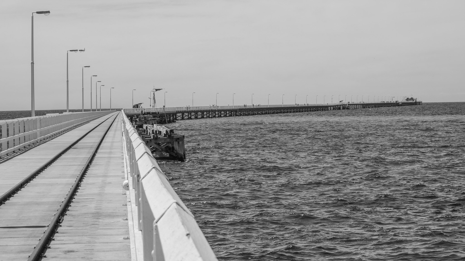 A long empty pier extends over the choppy ocean, featuring a railway track and railing, evoking themes of solitude and vastness.