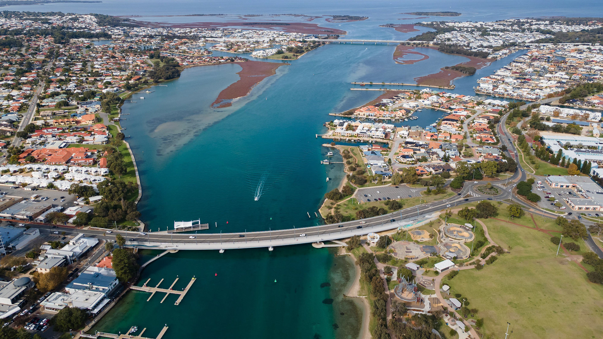 This stunning aerial stock image captures the breathtaking view of a coastal town that features a winding river, residential areas, and a marina. The vibrant turquoise waters contrast beautifully with the brown estuarine environment. Lush greenery surrounds the area, complemented by carefully arranged neighborhoods and infrastructure. The scene showcases a bridge connecting two parts of the town and highlights the serene lifestyle that accompanies living by the water. Ideal for use in travel articles, real estate brochures, and advertisements highlighting coastal living.