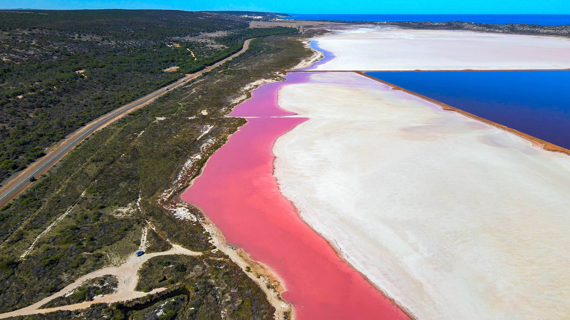 An aerial view reveals a striking pink salt lake beside deep blue water, showcasing unique geological formations and vibrant colors.