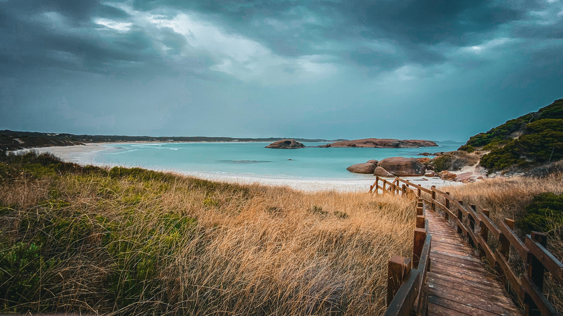 A wooden boardwalk winds through dry grass to a tranquil beach with turquoise waters, inviting exploration of nature's beauty.
