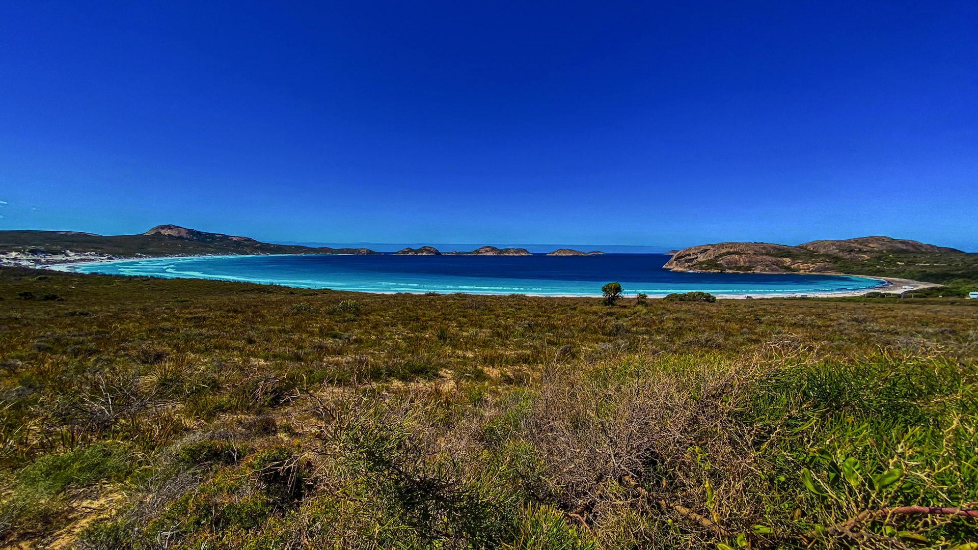 A serene coastal landscape showcases a vibrant ocean, sandy beach, and clear blue sky. Rugged hills and islands enhance the view.