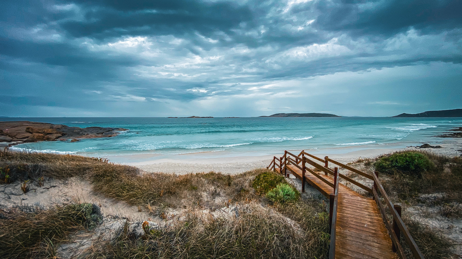 A wooden boardwalk descends to a tranquil beach, where ocean waves meet sand. The cloudy sky enhances the serene coastal landscape.