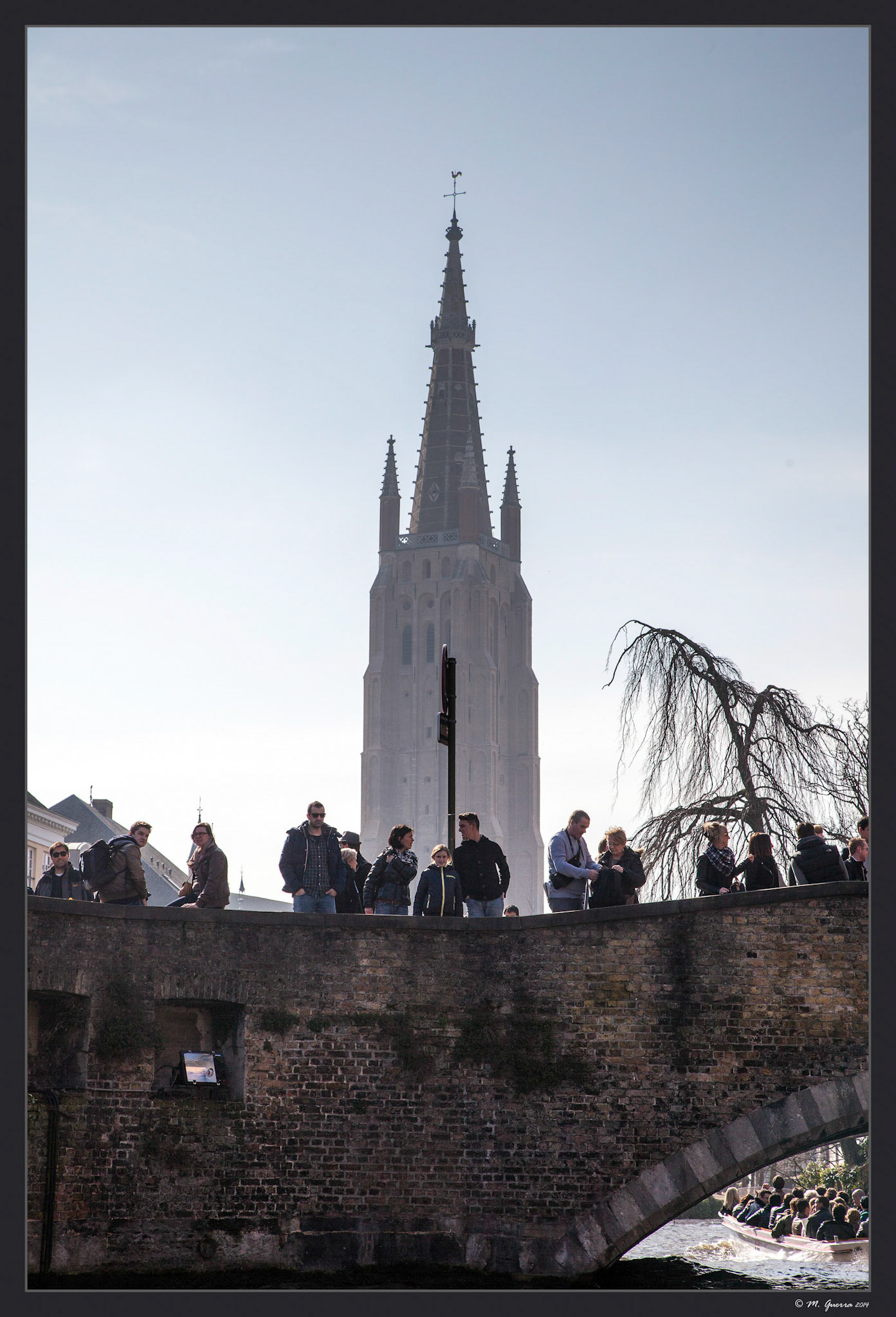 Turistas sobre un puente de Brujas con la torre de una iglesia al fondo. Ampliar