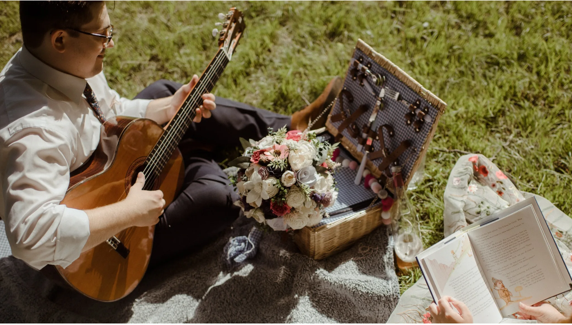 Isaac Brady performing an acoustic guitar on a picnic blanket in a field.