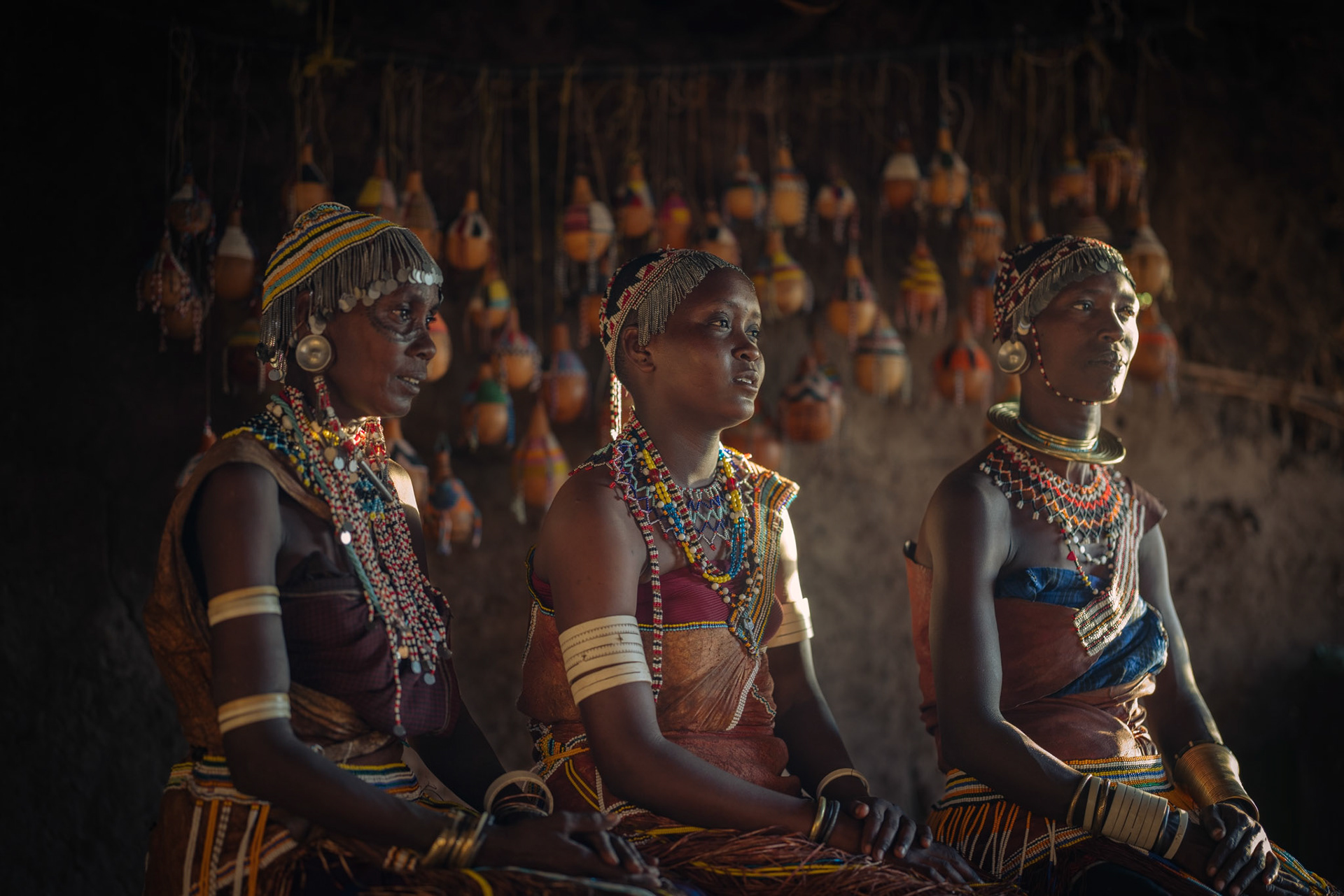 The Council of Women — Gathered within the cool shelter of the hut, three generations of women share a moment of connection. The play of light and shadow highlights the strong communal bonds that hold the tribe together, preserving wisdom and unity in a changing world.