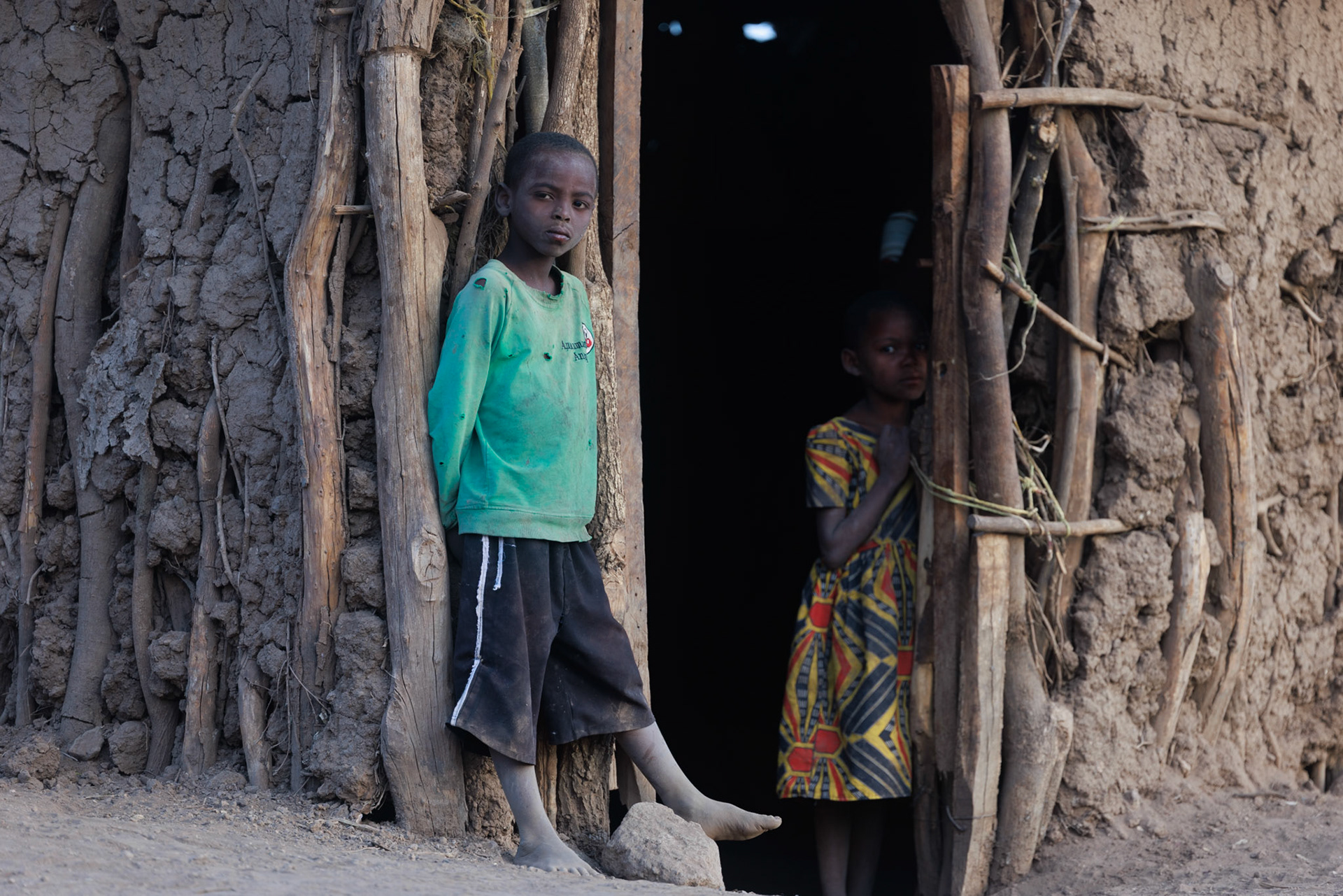 The Next Generation — Two Datoga children stand at the threshold of their family boma, their gaze reflecting a quiet curiosity. This image bridges the gap between ancient traditions and the future, capturing the innocence of youth within the rugged beauty of their ancestral home.