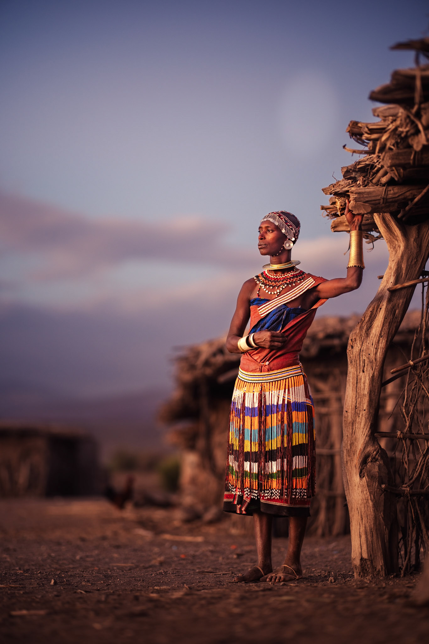 Pillar of the Boma — Leaning against the rough-hewn timber of her home, she wears traditional tanned leather garments and brass ornaments that signify her marital status. This image captures the harmonious relationship between the Datoga people and the natural materials they use to build their lives.
