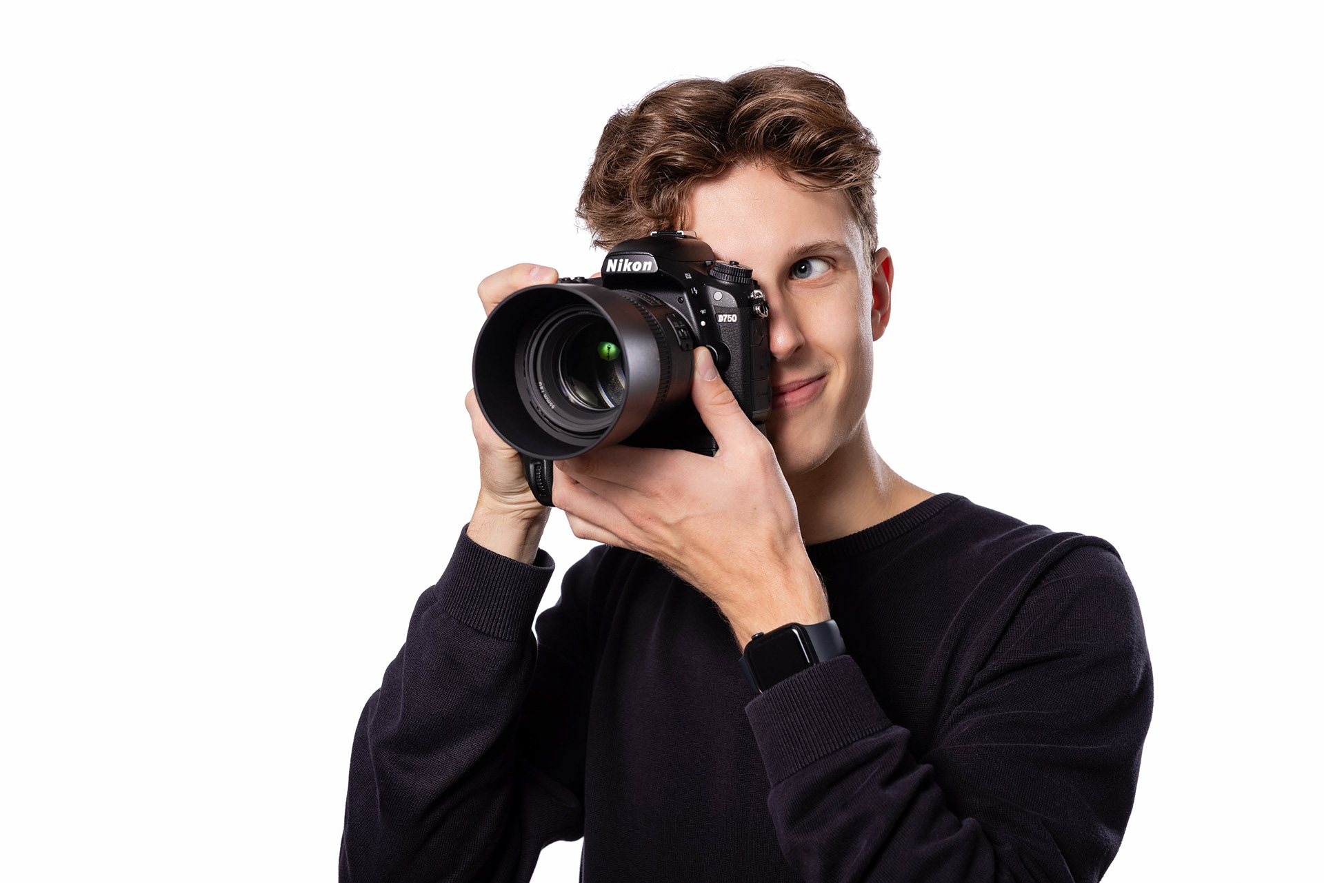 Junger Mann mit braunem Haar und Nikon-Kamera, konzentriert durch den Sucher schauend, vor weißem Hintergrund.