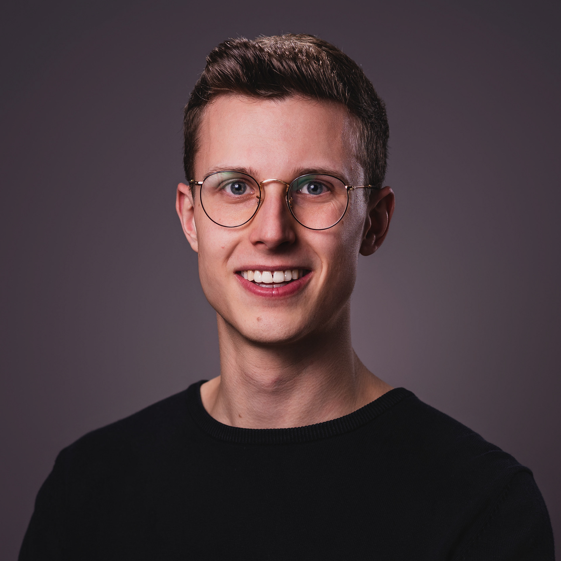 Ein junger Mann mit braunem, kurzem Haar trägt einen schwarzen Pullover und lächelt vor einem dunklen Hintergrund direkt in die Kamera.