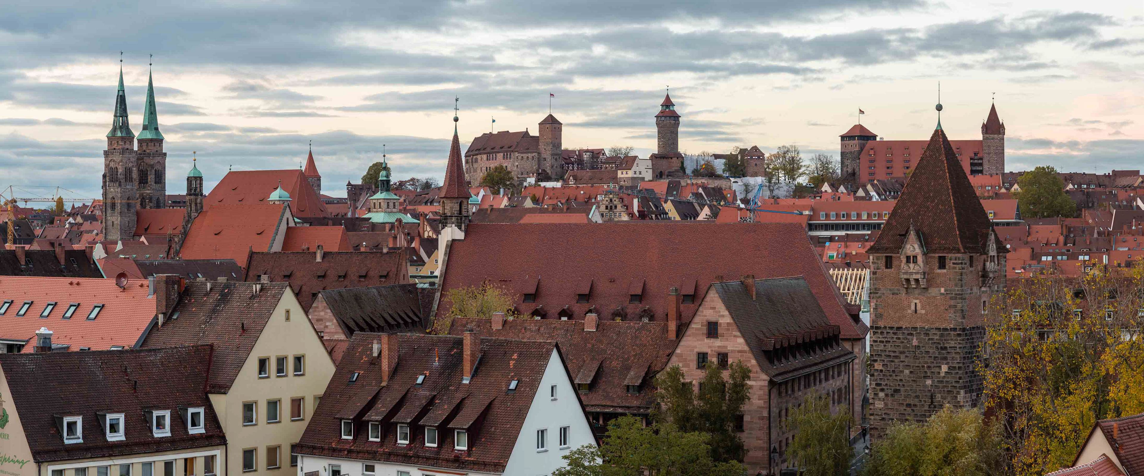 Blick auf die Skyline von Nürnberg mit historischen Gebäuden, roten Ziegeldächern, Türmen und der Kaiserburg im Hintergrund unter einem bewölkten Himmel.
