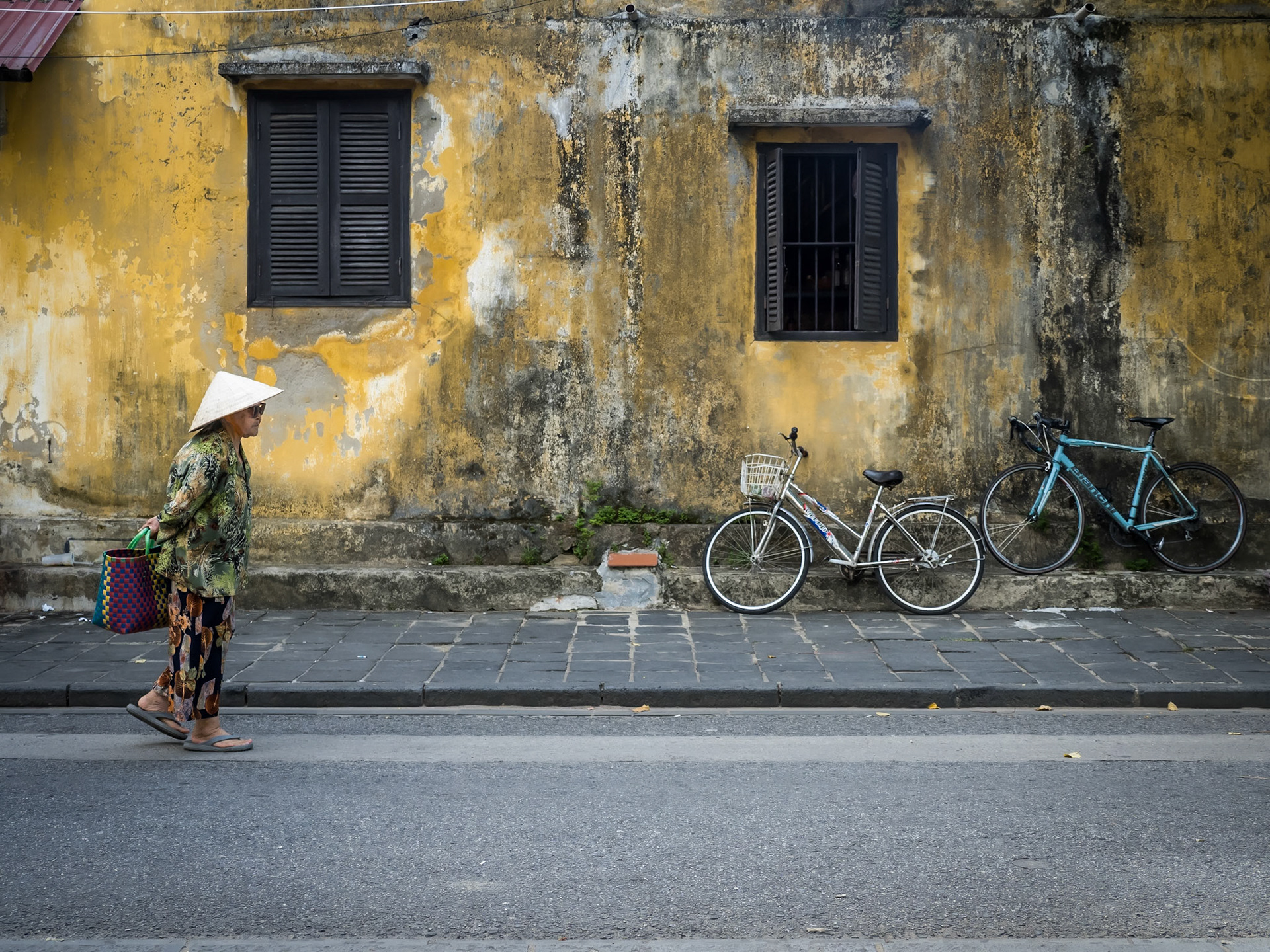 Against a beautifully weathered ochre wall, an elderly woman in a traditional Vietnamese 'leaf hat' walks past on her way to the local market