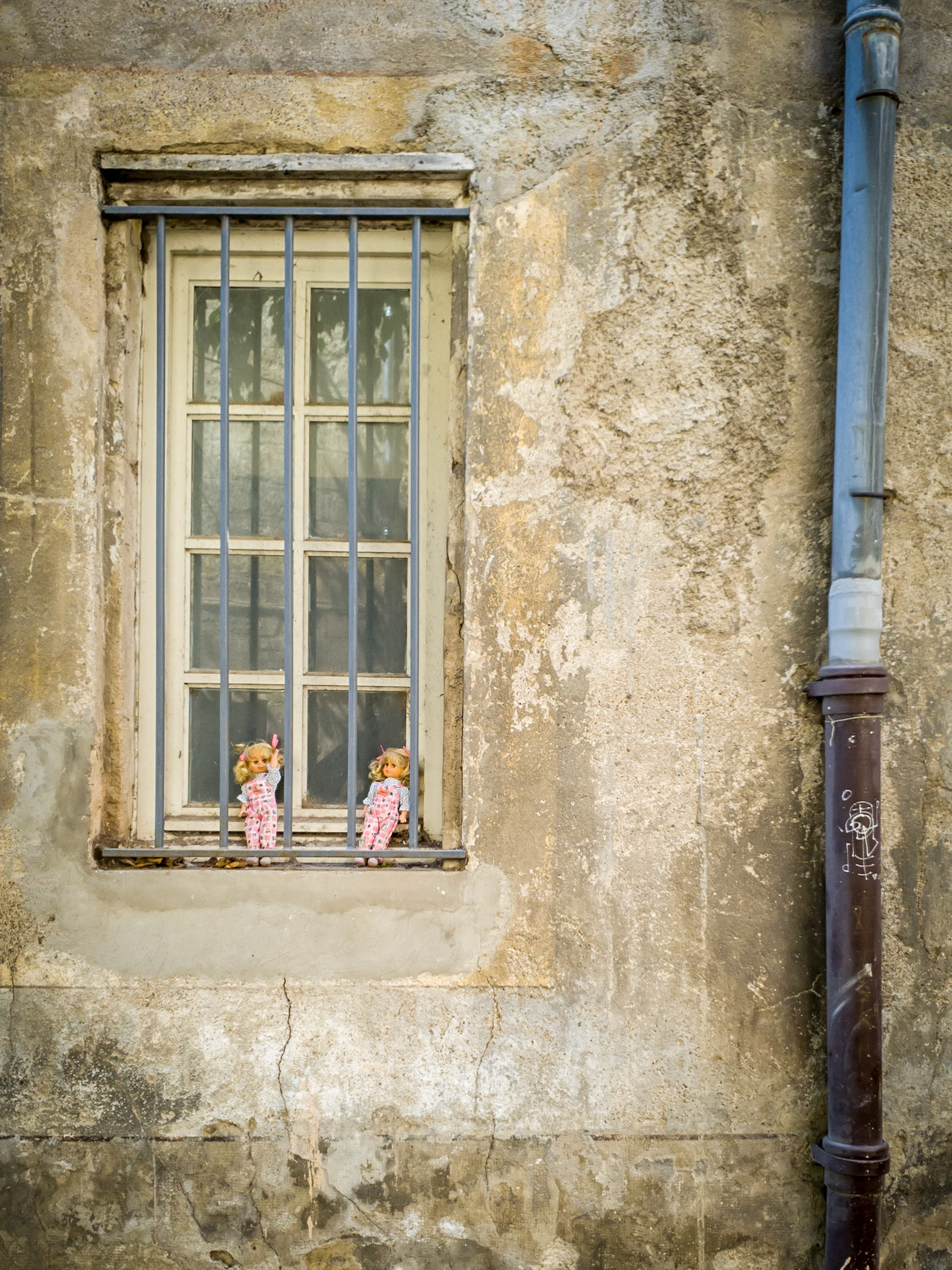 A pair of dolls sit on the barred windowsill of an old building in the Pezenas